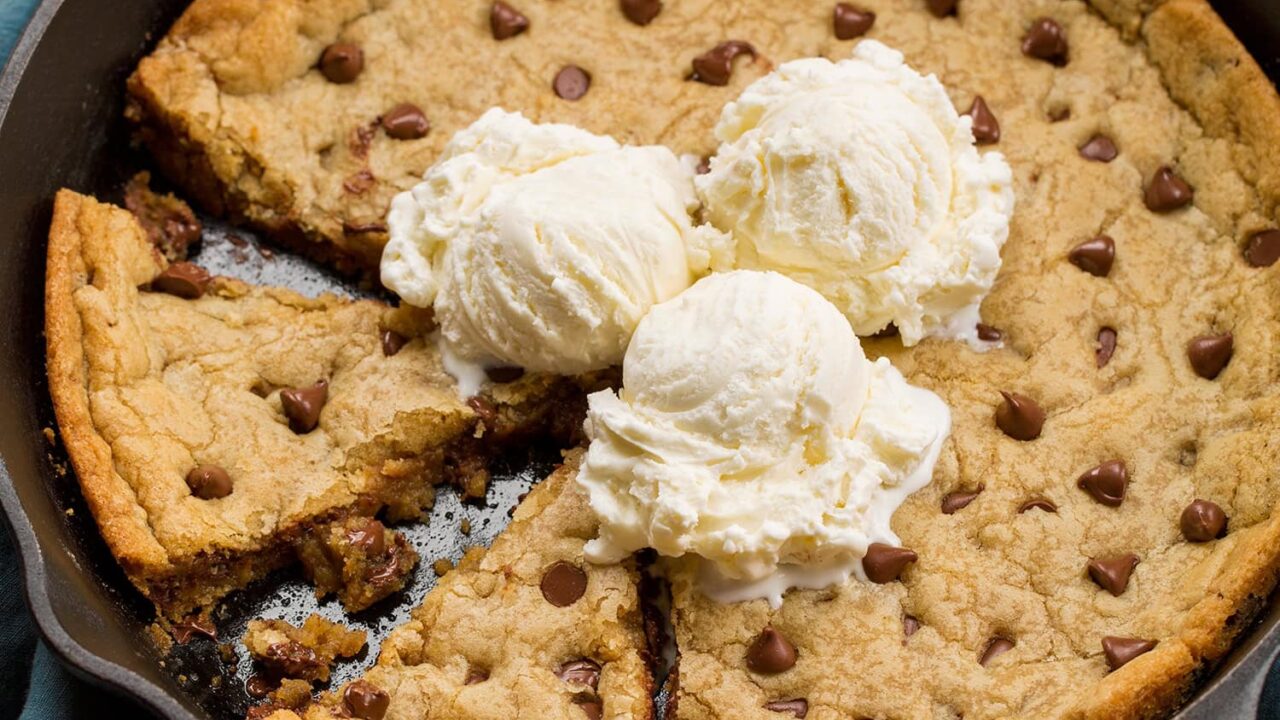Skillet chocolate chip cookie with two wedges cut and three vanilla ice cream scoops on top.